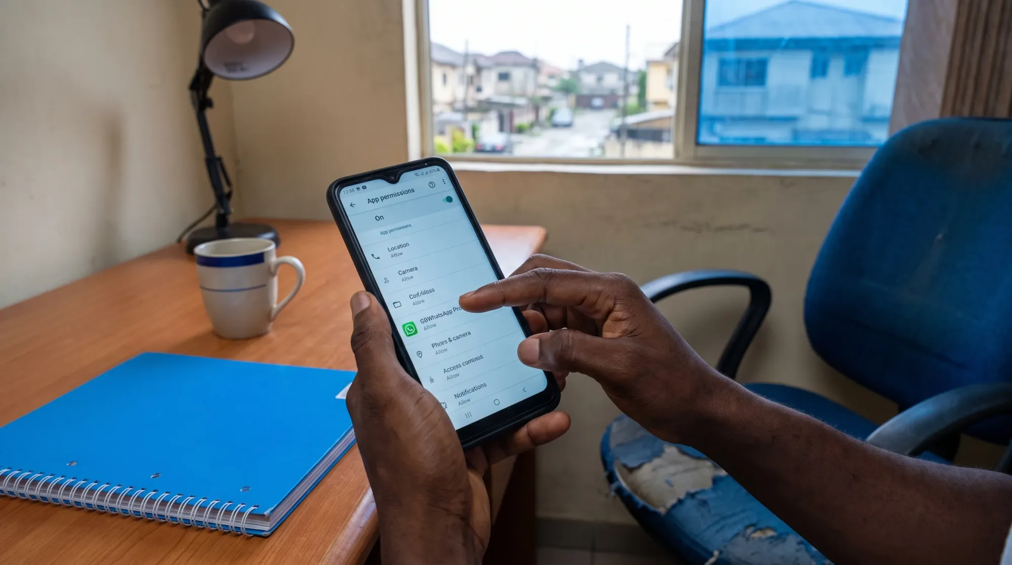 A Nigerian Android user checks app permissions at a simple home workspace before removing GBWhatsApp Pro, with the phone screen facing the camera and no private details visible.
