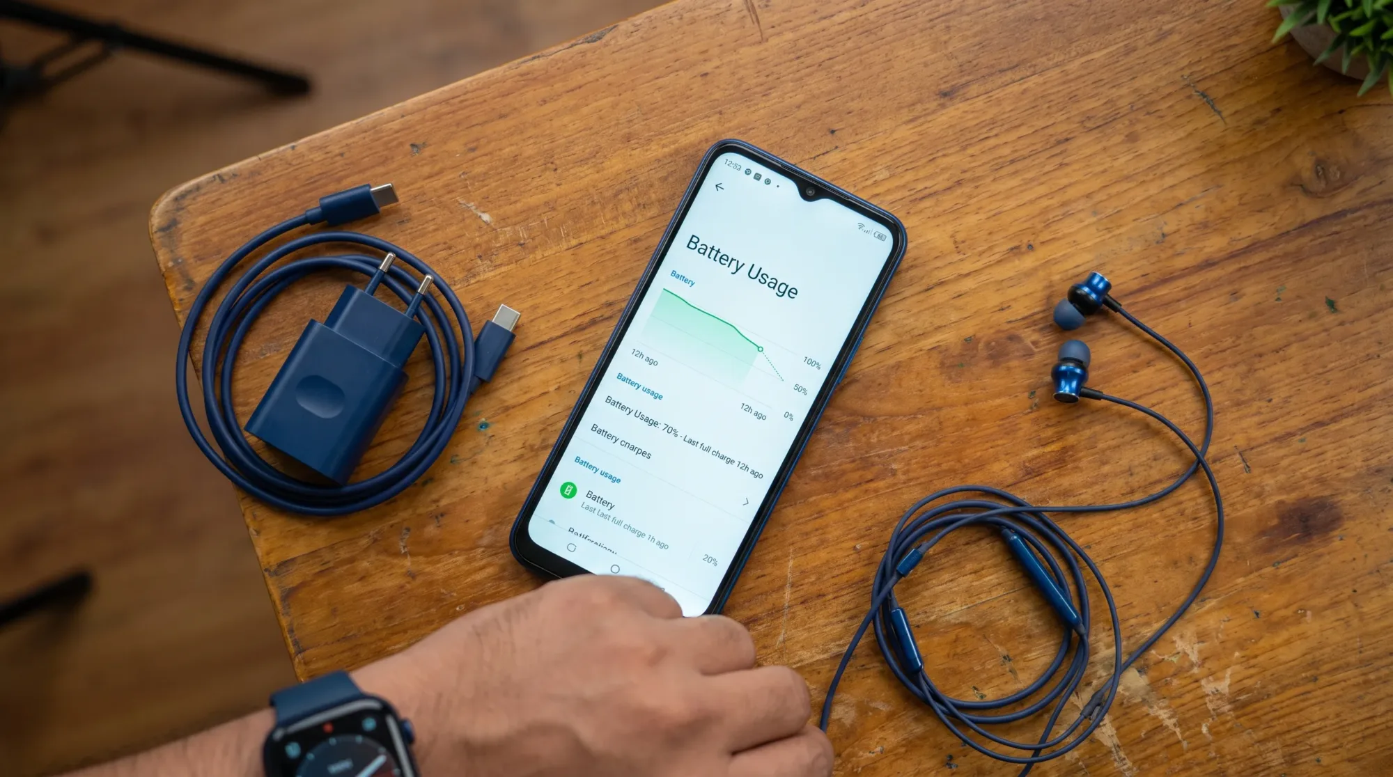 An Infinix Hot 12 Play lies on a wooden desk beside a charger and earphones while battery usage is checked on the screen.
