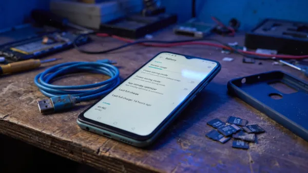 An Infinix Hot 12 Play sits on a repair bench beside a charging cable and memory cards, with battery settings open on the screen.