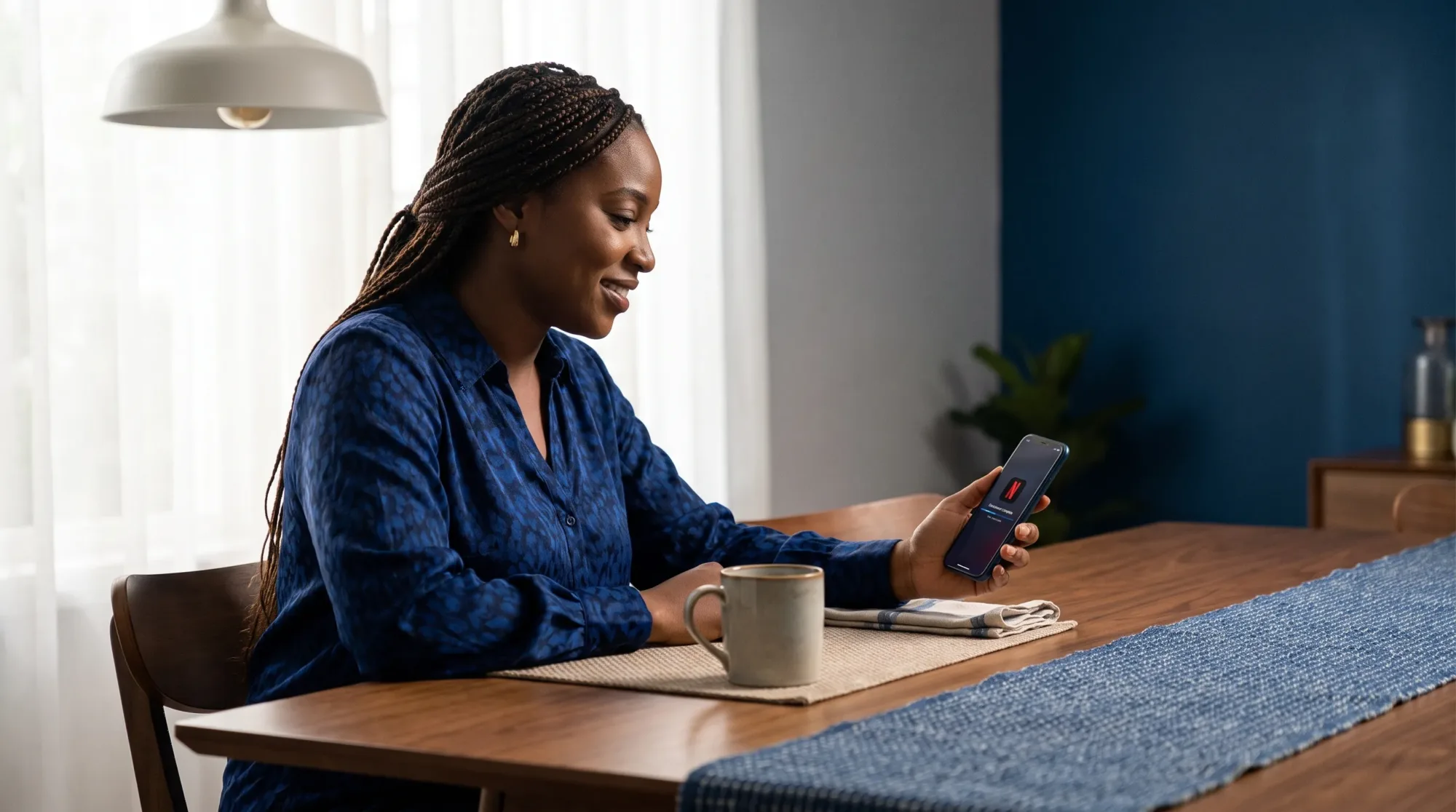 A Nigerian woman at a dining table checking her phone after a Netflix app download, with the screen angled toward her and no readable account details visible.